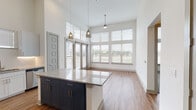 This single-floor residence is defined by its dramatic soaring ceilings, which lend an expansive, airy quality to the open-concept living and kitchen areas. Large windows with horizontal blinds line the living space, allowing abundant natural light to fill the interior while offering views of the surrounding greenery. Warm hardwood-style flooring runs throughout the main living areas, complementing the clean, contemporary design.

The kitchen serves as the centerpiece of the space, anchored by a substantial island with polished granite countertops and navy-blue cabinetry with brushed gold hardware. The perimeter cabinetry is finished in crisp white shaker-style panels, paired with a herringbone-patterned tile backsplash and under-cabinet lighting. Stainless steel appliances, including a refrigerator with ice maker, range, microwave, and dishwasher, complete the kitchen. Pendant lighting hangs above the island and over the adjacent dining area.

The bedroom features neutral carpet and connects directly to a private bathroom equipped with a soaking tub, marble-style tile surround, granite vanity, and an adjacent walk-in closet. A dedicated laundry area houses a full-size top-load washer and dryer. The overall ambiance is clean, modern, and functional throughout.
