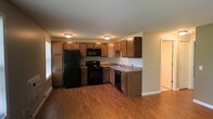 This single-floor space centers around a well-appointed kitchen that combines practicality with a warm aesthetic. The kitchen features medium-toned wood cabinetry arranged in an L-shaped layout, providing ample upper and lower storage throughout. Granite-style countertops offer generous prep space, complementing the full suite of black appliances, which includes a refrigerator, electric range, built-in microwave, and dishwasher. A double-basin sink is positioned beneath upper cabinets along the far wall, completing the functional layout. Rich hardwood flooring runs throughout, adding warmth and visual continuity to the space. Flush-mount ceiling fixtures provide consistent overhead lighting, keeping the kitchen bright and well-lit. A window along the left wall allows natural light to filter in, enhancing the overall brightness. The kitchen opens directly into an adjoining area, creating a connected, open feel across the single floor. A doorway visible at the far right suggests additional rooms beyond, offering a natural flow throughout the layout. The overall ambiance is clean, practical, and move-in ready.