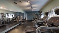 This fitness center is designed to provide a functional and motivating workout environment. The space features light teal walls accented with fitness-themed typography in bold lettering, adding an energetic and purposeful atmosphere. Track lighting runs along the textured ceiling, illuminating the room effectively and highlighting the equipment below.

Along one wall, floor-to-ceiling mirrors create a sense of expanded space and allow users to monitor their form during exercise. The cardio area is well-stocked with a row of treadmills and elliptical machines, including Star Trac branded equipment, positioned along the right side of the room. Strength training equipment, including weight benches and resistance machines, occupies the mirrored section of the space.

Dark rubber flooring provides durability and safety throughout the room, offering appropriate cushioning for both cardio and strength activities. A window along the back wall allows natural light to supplement the track lighting. The overall layout is efficient, maximizing usable floor space while maintaining clear pathways between equipment, creating an accessible and practical fitness facility.