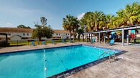 This outdoor amenity space in Mobile, Alabama, is centered around a generously sized swimming pool with clear blue water, lane dividers, and built-in entry steps with stainless steel handrails. The pool deck is finished in a stamped concrete surface and lined with blue lounge chairs, offering ample space for relaxation under the open sky. A black iron security fence encloses the pool area, providing both safety and a defined perimeter.

Adjacent to the pool, a pergola structure offers shaded gathering space, complemented by additional seating and a colorful umbrella-covered table nearby. Mature palm trees and lush greenery frame the area, enhancing the resort-like atmosphere. Landscaped beds with ornamental palms and mulched plantings add visual interest throughout the grounds.

A covered walkway runs alongside the brick-exterior building, with a concrete path leading from the covered porch area directly toward the pool. French doors with glass pane inserts serve as an entry point from the covered porch, which also houses a vending machine for added convenience. The surrounding grounds feature well-maintained grass and additional community buildings visible in the background, contributing to the cohesive, established character of this Mobile, AL community.