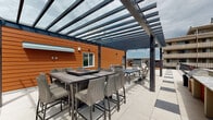 Located at 1122 Albion St in Denver's vibrant 80220 neighborhood, this rooftop terrace offers a well-appointed outdoor retreat with striking architectural detail and expansive city views. The terrace is anchored by a sleek steel-framed pergola with a translucent panel roof, allowing natural light to filter through while providing partial shade over the primary gathering area. Large-format porcelain tiles in a light cream and charcoal checkerboard pattern extend across the entire deck, lending a clean, contemporary aesthetic. The pergola shelters multiple high-top dining tables paired with woven rattan barstools, accommodating generous group seating. Additional seating areas beneath and beyond the pergola include swivel dining chairs and a separate long table suited for communal gatherings. Along the open perimeter, chaise lounge chairs invite sun-soaking, while potted greenery and a built-in outdoor kitchen station with an orange accent cabinet add functional and decorative character. The terrace is enclosed by a sleek dark metal railing, providing unobstructed panoramic views of the Denver urban landscape under Colorado's signature blue skies. The warm orange horizontal-panel cladding of the adjacent rooftop structure provides a bold, modern contrast to the neutral tones of the deck, completing this dynamic rooftop amenity space.