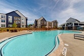 This multi-story residential community showcases a compelling blend of modern and transitional architectural design, characterized by bold navy blue and crisp white board-and-batten exterior finishes, steeply pitched gabled rooflines, and private balconies that overlook the grounds below. The development spans two floors of thoughtfully designed living spaces across several interconnected buildings.

At the heart of the community lies a generously sized freeform swimming pool, its clear turquoise waters serving as the centerpiece of an expansive outdoor amenity area. The surrounding pool deck is outfitted with numerous lounge chairs and market umbrella-shaded seating clusters, providing ample space for sunbathing and relaxation. The pool area is secured by a wrought iron perimeter fence, ensuring a private and safe environment.

Beyond the pool, the outdoor amenity space continues with a dedicated lawn area featuring a freestanding hammock, perfect for leisurely lounging. A charming pergola with string lights provides a shaded gathering spot, while additional patio seating areas invite casual outdoor socializing. Lush landscaping with young trees and manicured greenery adds to the welcoming atmosphere. The entire community is bathed in natural sunlight, with open skies contributing to its bright, airy, and resort-like ambiance.