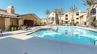 This outdoor amenity space is designed in a Mediterranean-inspired style, characterized by warm stucco exteriors, clay tile rooflines, and arched architectural details that lend the area a refined, resort-like atmosphere. The centerpiece is a generously sized swimming pool with clear blue water, surrounded by a broad concrete pool deck ideal for lounging. Neatly arranged chaise lounges line the perimeter, offering dedicated sunbathing areas beneath the open sky.

Flanking the pool area are elegantly appointed cabanas featuring dark metal frames and rich burgundy and striped fabric curtains that can be drawn for privacy or shade. Inside each cabana, dark wicker sectional sofas and ottomans with cream cushions provide comfortable seating for relaxation or socializing. Tall palm trees and strategically placed planters with ornamental succulents and tropical plants add lush greenery throughout the space.

Adjacent to the pool deck, an outdoor dining area includes table and chair sets positioned near what appears to be a barbecue station, making the space functional for entertaining. Wrought-iron fencing borders sections of the courtyard, maintaining an open yet defined layout. Overall, the space conveys a well-curated, upscale outdoor living environment suited for leisure and social gatherings.