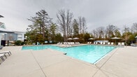Located at 9015 Kings Grant Dr in Charlotte, North Carolina, this community amenity space centers around a generously sized outdoor swimming pool with crystal-clear blue water. The pool deck is finished in smooth concrete and outfitted with multiple lounge chairs and shaded umbrella stations, providing ample space for relaxation and recreation. A black wrought-iron fence encloses the pool area, enhancing both safety and visual appeal, while tall mature trees in the background create a natural, private backdrop.

Adjacent to the pool is a well-maintained amenity building constructed with warm brick masonry and complemented by light blue lap siding — a combination that gives the space a welcoming, residential character. The building features white-framed windows, lantern-style exterior lighting, and glass-panel entry doors that connect the indoor and outdoor areas seamlessly. A paved patio area along the building's exterior provides additional seating with lounge chairs and a stylish navy-patterned umbrella. Potted flowering plants add decorative touches near the entryway. Security cameras and exterior flood lighting are also present, reflecting a commitment to the safety and comfort of residents. The overall ambiance is clean, functional, and inviting — ideal for outdoor leisure in the Charlotte community.