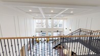 This impressive space makes an immediate statement with its grand, open lobby anchored by a sweeping staircase with dark wrought-iron balusters and a warm wood handrail. The soaring double-height ceiling is adorned with a refined coffered design accented by recessed lighting, lending a sense of architectural sophistication throughout. Crisp white wainscoting panels line the walls, contributing to a clean, classic aesthetic that feels both welcoming and polished.

Large, multi-pane windows along the front facade flood the main lobby with natural light, offering views of the surrounding streetscape. The floor features a bold geometric tile pattern in contrasting tones, adding visual interest at ground level. Seating areas are thoughtfully arranged throughout, including vibrant accent chairs in yellow near the main entrance and plush purple lounge chairs positioned beside oversized windows that look out onto a landscaped courtyard with stone ground cover and views of the adjacent building.

The overall ambiance of the space balances traditional architectural detailing with contemporary furnishings, creating an inviting and functional environment. The thoughtful use of natural light, layered finishes, and carefully curated seating areas establishes a warm, welcoming atmosphere suited to a well-appointed residential or hospitality setting.