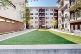 Located at 385 N Angier Ave NE in Atlanta's Old Fourth Ward neighborhood, this outdoor courtyard space serves as a central amenity area nestled within a modern multi-story residential complex. The courtyard is thoughtfully designed to balance greenery, hardscape, and communal gathering areas into a cohesive and inviting outdoor environment.

At the heart of the space lies a generous expanse of lush artificial turf lawn, framed by corten steel edging that provides a clean, contemporary boundary. Wide, grid-patterned concrete paver walkways run alongside the lawn, creating clearly defined pathways that connect various areas of the courtyard. String lights are strung overhead, adding a warm, ambient glow during evening hours.

Raised planting beds line the perimeter, filled with a variety of greenery, flowering plants, and what appears to be a dedicated garden area with cultivated raised beds — ideal for community gardening. Mature trees are interspersed throughout, providing natural shade and a sense of established character.

Communal seating areas are thoughtfully positioned across the courtyard, including an umbrella-shaded picnic table and casual lounge furniture, encouraging social interaction and relaxation. The surrounding multi-story building features private balconies that overlook the courtyard, integrating the outdoor space with the residences above. The overall ambiance is open, bright, and welcoming, with abundant natural light throughout the day.
