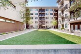 Located at 385 N Angier Ave NE in Atlanta's Old Fourth Ward neighborhood, this outdoor courtyard space serves as a central amenity area nestled within a modern multi-story residential complex. The courtyard is thoughtfully designed to balance greenery, hardscape, and communal gathering areas into a cohesive and inviting outdoor environment.

At the heart of the space lies a generous expanse of lush artificial turf lawn, framed by corten steel edging that provides a clean, contemporary boundary. Wide, grid-patterned concrete paver walkways run alongside the lawn, creating clearly defined pathways that connect various areas of the courtyard. String lights are strung overhead, adding a warm, ambient glow during evening hours.

Raised planting beds line the perimeter, filled with a variety of greenery, flowering plants, and what appears to be a dedicated garden area with cultivated raised beds — ideal for community gardening. Mature trees are interspersed throughout, providing natural shade and a sense of established character.

Communal seating areas are thoughtfully positioned across the courtyard, including an umbrella-shaded picnic table and casual lounge furniture, encouraging social interaction and relaxation. The surrounding multi-story building features private balconies that overlook the courtyard, integrating the outdoor space with the residences above. The overall ambiance is open, bright, and welcoming, with abundant natural light throughout the day.