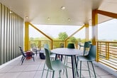 Located at 715 E 36th St in Charlotte, NC, this covered rooftop terrace offers a compelling blend of modern design and open-air comfort. The space is defined by its striking yellow structural beams and diagonal bracing, which frame panoramic views of the surrounding tree canopy and Charlotte skyline. A clean white ceiling with recessed lighting overhead provides shelter while maintaining a bright, airy atmosphere throughout.

The terrace is finished with large-format light gray floor tiles, lending a polished yet casual aesthetic underfoot. The layout accommodates multiple seating configurations, including a high-top bar table surrounded by contemporary green stools, a lounge corner with woven rattan chairs and accent side tables, and a dining area featuring a slatted green table with coordinating bench seating — each zone designed to support different types of social interaction.

One interior wall is clad in vertical sage-green metal siding, adding a textural architectural element and anchoring the space visually. A wall-mounted screen is also present, extending the terrace's functionality for events or presentations. The open-railing perimeter allows unobstructed sightlines to the lush greenery below, making this an inviting and thoughtfully designed communal outdoor amenity.