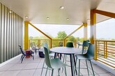 Located at 715 E 36th St in Charlotte, NC, this covered rooftop terrace offers a compelling blend of modern design and open-air comfort. The space is defined by its striking yellow structural beams and diagonal bracing, which frame panoramic views of the surrounding tree canopy and Charlotte skyline. A clean white ceiling with recessed lighting overhead provides shelter while maintaining a bright, airy atmosphere throughout.

The terrace is finished with large-format light gray floor tiles, lending a polished yet casual aesthetic underfoot. The layout accommodates multiple seating configurations, including a high-top bar table surrounded by contemporary green stools, a lounge corner with woven rattan chairs and accent side tables, and a dining area featuring a slatted green table with coordinating bench seating — each zone designed to support different types of social interaction.

One interior wall is clad in vertical sage-green metal siding, adding a textural architectural element and anchoring the space visually. A wall-mounted screen is also present, extending the terrace's functionality for events or presentations. The open-railing perimeter allows unobstructed sightlines to the lush greenery below, making this an inviting and thoughtfully designed communal outdoor amenity.