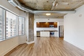 Located at 4501 Ford Ave in Alexandria, VA, this one-bedroom, two-bathroom residence blends industrial character with modern finishes across a single thoughtfully designed floor. The open-concept living, dining, and kitchen area serves as the heart of the home, highlighted by exposed concrete columns, visible steel ductwork, and light wood-plank flooring that unifies the space. A striking wall of floor-to-ceiling windows floods the living area with abundant natural light while offering panoramic views of the surrounding Alexandria skyline. The kitchen features rich walnut-toned upper cabinetry, white lower cabinets, dark countertops, and a full suite of stainless steel appliances, including a refrigerator, range, and dishwasher. A central hallway connects the private quarters to the main living area, lined with louvered closet doors for convenient storage access. The generously sized primary bedroom features the same wood-plank flooring and an exposed concrete ceiling, adding to its loft-like aesthetic. The en-suite bathroom is finished with large-format tile flooring, a white quartz vanity with gray cabinetry, and a glass-enclosed shower. A walk-in closet with built-in shelving and hanging rails provides ample storage. A dedicated laundry room and utility room add further practicality to this distinctive urban residence.