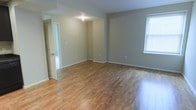 This single-floor space presents a clean and functional layout with a welcoming, move-in-ready aesthetic. The main living area is characterized by warm hardwood-style flooring that runs throughout, lending a sense of continuity and warmth to the interior. Neutral gray-toned walls provide a versatile backdrop, complementing a variety of décor styles and color schemes.

Natural light filters in through a window fitted with blinds, brightening the open living area and highlighting the polished surface of the floors. The space benefits from a practical, open-concept feel, with clearly defined zones that allow for flexible furniture arrangements. Recessed and overhead lighting supplements the natural light, ensuring the interior remains well-lit throughout the day.

An interior doorway leads to an adjacent room or hallway, suggesting a thoughtful flow between spaces. White baseboards and clean wall edges add a crisp, finished look to the overall interior. The absence of clutter and the simplicity of the design contribute to an atmosphere that feels airy and unconfined. Overall, this space offers a straightforward, well-maintained environment with solid foundational finishes suited to a range of uses and lifestyles.