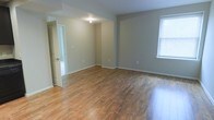 This single-floor space presents a clean and functional layout with a welcoming, move-in-ready aesthetic. The main living area is characterized by warm hardwood-style flooring that runs throughout, lending a sense of continuity and warmth to the interior. Neutral gray-toned walls provide a versatile backdrop, complementing a variety of décor styles and color schemes.

Natural light filters in through a window fitted with blinds, brightening the open living area and highlighting the polished surface of the floors. The space benefits from a practical, open-concept feel, with clearly defined zones that allow for flexible furniture arrangements. Recessed and overhead lighting supplements the natural light, ensuring the interior remains well-lit throughout the day.

An interior doorway leads to an adjacent room or hallway, suggesting a thoughtful flow between spaces. White baseboards and clean wall edges add a crisp, finished look to the overall interior. The absence of clutter and the simplicity of the design contribute to an atmosphere that feels airy and unconfined. Overall, this space offers a straightforward, well-maintained environment with solid foundational finishes suited to a range of uses and lifestyles.