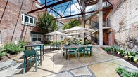 Located at 111 N Marietta Pkwy NE in Marietta, Georgia, this outdoor courtyard is a masterfully reimagined industrial space that blends historic architectural character with modern amenities. Enclosed by the weathered brick and corrugated metal facades of the surrounding building complex, the courtyard is anchored by an exposed steel overhead framework that frames open sky views, flooding the space with natural light throughout the day.

The grounds are divided into distinct functional zones. A generously sized dining area features multiple table-and-chair sets with large shade umbrellas, ideal for al fresco gatherings. Adjacent to this, a relaxed lounge area centers around a rectangular Corten steel fire pit, flanked by woven lounge chairs and benches, perfect for evening socializing. A dedicated grilling station with multiple gas grills is set along a beautifully crafted flagstone path bordered by lush, curated landscaping.

Mature trees, Japanese maples, tropical foliage, and layered plantings bring vibrant greenery throughout, softening the raw industrial backdrop. Concrete columns support open-riser staircases with black metal railings, adding verticality and visual interest. The mixed paving — combining large concrete slabs, mosaic flagstone inlays, and gravel accents — creates a thoughtfully designed hardscape that ties the entire courtyard together into a cohesive, inviting outdoor environment.