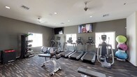 This fitness center offers a comprehensive workout environment spread across a single floor, thoughtfully organized to accommodate a variety of exercise routines. The space is finished with durable, multi-toned striped carpet flooring that defines the workout zones while adding a contemporary aesthetic. Neutral gray and white walls contribute to a clean, focused atmosphere, complemented by recessed lighting and ceiling fans that ensure comfort during workouts.

The cardio section is well-stocked with multiple treadmills, elliptical trainers, and a stair climber, each equipped with individual digital displays. Wall-mounted flat-screen televisions are positioned throughout, providing entertainment during cardio sessions. Natural light filters in through several windows fitted with blinds, brightening the space without causing glare on screens or equipment.

A dedicated strength training area houses a multi-station cable machine, a full dumbbell rack, adjustable weight benches, and exercise balls, offering versatile options for resistance training. A large mirror panel visually expands the space and allows users to monitor their form. An open passageway connects to an adjacent lounge or common area, adding convenience and accessibility to the overall facility.
