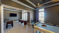This single-floor game and activity room is designed for both recreation and productivity, offering a diverse range of amenities within a cohesive, stylish environment. The space is defined by its sophisticated gray-toned walls, crisp white crown molding, and elegant tray ceilings with recessed lighting, all of which lend a polished and upscale ambiance throughout.

The main game area serves as the centerpiece of the space, featuring a striking white billiards table with ornate legs, a full-length shuffleboard table, and a foosball table — providing ample entertainment options. A wall-mounted flat-screen television anchors one end of the room, making it well-suited for watching sports or gaming. Light wood-plank flooring flows continuously throughout, adding warmth and cohesion.

Natural light pours in through multiple large windows, some framed with transom panels, offering views of the surrounding outdoor environment. French-style glass doors further enhance the connection between the interior and exterior. A dedicated workspace area equipped with desks and office chairs provides a quieter zone for productivity.

The layout is open and generously proportioned, allowing for comfortable movement between activity zones. The overall design strikes a balance between a lively recreational atmosphere and a refined, well-finished aesthetic, making this a versatile and inviting community amenity space.