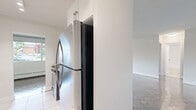 Located at 2292 Weston Rd in Toronto's Weston neighbourhood, this one-bedroom, one-bathroom unit offers a practical and well-organized layout across a single floor. The living room is a standout feature, highlighted by a wide bank of floor-to-ceiling windows fitted with roller blinds that flood the space with natural light while offering views of the surrounding residential streetscape. Baseboard heating units run beneath the windows, ensuring comfort year-round. The parquet wood flooring, found throughout the main living areas and bedroom, adds a classic character to the space. The living room flows into a separate kitchen area, which is visible through an open passageway and includes standard appliances. The bedroom is a comfortable, well-proportioned room featuring its own window for natural light, a built-in sliding-door closet for storage, and the same parquet flooring. The bathroom is finished in a clean, bright palette with large white wall tiles surrounding the bathtub and shower, light tile flooring, a toilet, and a dark-finish vanity with an integrated mirror. Neutral white walls throughout the unit provide a versatile backdrop, making the space easy to personalize.