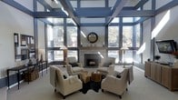 This refined lounge space makes an immediate impression with its dramatic architectural design. The standout feature is the bold coffered ceiling, finished in deep steel blue with integrated skylights that flood the interior with abundant natural light. Floor-to-ceiling windows line the walls, framed with sleek blinds that allow for adjustable light control while offering open views of the surrounding greenery outdoors.

At the heart of the room, a clean-lined stone fireplace serves as the focal point, accented by a wood mantel and a decorative sunburst mirror above. Flanking the fireplace, candle holders and understated décor maintain an elegant yet welcoming atmosphere. The seating arrangement — a curated circle of plush cream armchairs paired with natural wood stump side tables and anchored by a cowhide rug — encourages intimate conversation and relaxed gathering.

Warm floor lamps with classic shades provide soft ambient lighting that complements the natural brightness streaming in from the expansive windows. The neutral color palette of creams, grays, and warm wood tones is thoughtfully balanced against the bold architectural elements above. The overall ambiance is one of modern sophistication, offering a bright, airy, and inviting environment well-suited for social interaction and relaxation.
