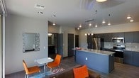 This single-floor residence showcases a thoughtfully designed open-concept layout that integrates the kitchen, dining, and living areas into one cohesive space. The kitchen is a standout feature, equipped with dark cabinetry, granite countertops, a double stainless steel sink, and a stainless steel refrigerator. Pendant lighting with glass fixtures hangs above the kitchen island, which doubles as a casual dining or entertaining surface, adding both function and visual interest.

The living area extends naturally from the kitchen, offering a comfortable and modern atmosphere. A sliding glass door provides direct access to an outdoor balcony, allowing natural light to fill the space and offering views of the surrounding area. Recessed lighting and track lighting complement the contemporary aesthetic throughout.

Dark hardwood-style flooring anchors the living space, where a cozy sofa with vibrant orange accent pillows and a compact bistro table with matching chairs add a modern, urban feel. A decorative mirror on one wall adds depth to the room. Multiple interior doors lead to additional private areas, including what appears to be a bathroom, suggesting a well-organized floor plan that balances open living with private spaces.