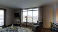 This single-floor residence centers around a thoughtfully designed living room that immediately draws attention to its large, floor-to-ceiling picture window, which frames an elevated view of the surrounding urban landscape. Natural light floods the space through the wide window fitted with horizontal blinds and framed by elegant patterned drape panels in neutral tones with bold geometric trim, adding a refined decorative touch.

The living room is finished with dark hardwood-style flooring that anchors the space and complements the warm, neutral wall tones. A patterned area rug in soft blues and creams defines the seating area, which includes a plush gray accent chair with a classic nail-head silhouette and a coordinating plaid throw. A mirrored console table along one wall provides both storage and a reflective design element, paired with decorative accessories and a wall-mounted flat-screen television. A traditional-style floor lamp adds ambient warmth to the room. The overall ambiance is polished and comfortable, blending classic furnishing styles with modern conveniences in a light-filled, elevated setting.