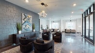 The lobby at 2303 Lebanon Pike in Nashville, Tennessee presents a contemporary and well-appointed first impression. Polished concrete floors extend throughout the open, single-level space, reflecting light and lending an industrial-modern aesthetic that ties the entire area together.

The main lounge area is anchored by a striking gray stone accent wall, against which a bold, colorful graffiti-style artwork commands attention. A geometric cube pendant light fixture serves as a focal design element overhead. Dark, curved upholstered lounge chairs are arranged on a patterned area rug, creating an inviting seating cluster. Tall potted plants punctuate the space with natural greenery. Deeper into the lounge, a secondary seating arrangement with lighter furnishings and tall pendant lighting benefits from abundant natural light streaming through large windows dressed in sheer white curtains.

The entry vestibule features black-framed glass double doors with transom windows above, contributing to the clean, modern architectural language of the space. A dedicated leasing office area is set apart with its own polished concrete flooring, pendant lighting, sheer-draped windows, and a dark executive desk flanked by neutral upholstered guest chairs — functional yet refined. A herringbone-patterned credenza adds a warm, textured detail to the office setting. Together, these areas create a cohesive, stylish, and highly functional lobby environment.