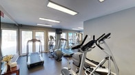 This fitness center is a thoughtfully designed, single-floor exercise facility that offers a comprehensive workout environment. The space is finished with durable, dark rubberized flooring throughout, providing a practical and resilient surface suited for a variety of fitness activities.

Natural light floods the room through a series of large windows along the exterior walls, offering views of the outdoor surroundings, including what appears to be a pool area and landscaped grounds. Recessed rectangular panel lighting supplements the natural light, ensuring the space remains bright and well-lit at all times.

The walls are painted in a cool, neutral grey tone, lending the space a clean and modern aesthetic. A large wall-mounted mirror on one side visually expands the room and allows users to monitor their form during workouts.

The facility is well-stocked with cardio equipment, including multiple treadmills, elliptical trainers, and a stationary bike. A wall-mounted multi-function cable and weight machine anchors the strength training area, offering a range of resistance exercises. A mounted flat-screen television is positioned for viewing during workouts. An open stretch of wall provides additional space for floor-based exercises or stretching. The fitness center connects to an adjacent hallway, offering easy access to the broader facility.
