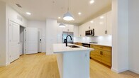 Located at 19320 US Highway 19 N in Clearwater, Florida, this single-floor unit showcases a clean, modern design throughout. The kitchen serves as a central highlight, featuring a two-tone cabinet scheme that pairs crisp white upper cabinetry with warm natural wood lower cabinets, all accented by matte black hardware. A large center island with a white quartz countertop and an undermount sink with a matte black faucet anchors the space, illuminated by stylish pendant lights overhead. Stainless steel appliances, including a refrigerator with an ice and water dispenser, a range, and an over-the-range microwave, complete the well-appointed kitchen. Light hardwood flooring flows consistently throughout the unit, lending warmth and visual continuity. The living area benefits from natural light streaming through large windows fitted with horizontal blinds, and a ceiling fan promotes comfortable airflow. Additional rooms are bright and open, with neutral white walls and ample natural light from generously sized windows. The bathroom features a wood-toned vanity with a white countertop, matte black hardware, a tub-shower combination with white subway tile surround, and coordinating light wood flooring, maintaining the unit's cohesive contemporary aesthetic.