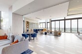 Located at 3201 N Federal Highway in Oakland Park, Florida, this sophisticated amenity lounge spans nearly 3,000 square feet of thoughtfully designed communal space on a single floor. The open-plan layout is anchored by large floor-to-ceiling windows that wrap around the perimeter, flooding the interior with abundant natural light and framing sweeping panoramic views of the South Florida skyline and surrounding landscape.

The space is divided into several distinct functional zones that flow seamlessly into one another. A generous lounge area features multiple seating arrangements, including plush modular sofas, swivel accent chairs, and coffee tables arranged atop vibrant blue area rugs — creating an inviting environment for relaxation and socializing. A dedicated game room zone includes a full-size pool table and a long dining-height game table with blue upholstered chairs, set beneath a striking curved ceiling detail with integrated gold lighting.

A well-appointed bar and kitchen area showcases a bold blue island with bar seating, a decorative tile backsplash, open wood shelving, and a white marble-topped island. Warm wood-toned columns, built-in shelving with sage green accents, and light-toned large-format tile flooring with geometric inlay patterns contribute to the space's refined, contemporary aesthetic throughout.