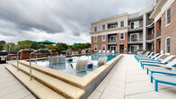 Located at 5820 Patterson Ave in Richmond, Virginia, this outdoor amenity space is centered around a generously sized resort-style swimming pool with pristine turquoise water and a classic tile surround. The pool deck is finished in large-format light gray pavers, providing ample space for lounging and socializing. White and blue chaise lounges are arranged along the pool's perimeter, offering plenty of spots for relaxation and sun exposure. A shallow wading ledge with in-water lounge chairs adds a distinctive touch, ideal for leisure in a shallow, comfortable setting. Accessibility features, including a pool lift and entry handrails, are also present. The deck extends into a dedicated outdoor entertaining area equipped with dining tables, chairs, and green market umbrellas, as well as grilling stations along the perimeter fencing. The space is enclosed on multiple sides by a multi-story brick and stucco residential building with private balconies overlooking the pool, creating a courtyard-like atmosphere. Surrounding treetops are visible beyond the low perimeter railing, contributing to the sense of an open, airy outdoor retreat in an established Richmond neighborhood.