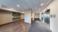 The lobby of Vibe Apartments at Arbour Lake in Calgary, AB, serves as an inviting and well-appointed entry point for residents and visitors alike. The space features a contemporary design with a combination of warm hardwood-style flooring and durable carpeted pathways that guide movement through the area. A prominent feature wall displays the "Vibe Apartments at Arbour Lake" branding, accented by a lush vertical greenery installation set against a stylish textured grey backdrop, creating a striking first impression upon entry. The lobby includes a comfortable lounge area furnished with two upholstered armchairs, a round coffee table, and a tropical potted plant, all positioned beside a window that brings in natural light. Warm wood slat paneling adds an architectural accent nearby. A built-in counter with bar-style seating provides a functional workspace area. Practical amenities are well-integrated, including a bank of resident mailboxes, a floor directory panel, a resident intercom system near the entrance, and a stainless steel elevator with clearly marked floor signage. Recessed ceiling lighting throughout ensures the space is bright and welcoming, while clean white walls and neutral tones contribute to the overall modern and polished ambiance of this residential lobby.