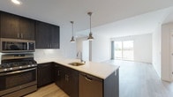 This single-floor residence showcases a thoughtfully designed open-concept layout that seamlessly connects the kitchen and living areas. The kitchen is a standout feature, equipped with dark espresso-toned cabinetry, a white quartz peninsula with an undermount sink, stainless steel appliances including a gas range, microwave, refrigerator, and dishwasher, complemented by a white subway tile backsplash. Two industrial-style pendant lights hang above the peninsula, adding a modern accent.

The living area is bright and airy, benefiting from natural light streaming through a large sliding glass door that opens onto a private balcony with outdoor views. Light wood-tone flooring runs throughout the main living spaces, creating a warm, cohesive aesthetic.

The space includes two bathrooms, each distinctly designed. One features dark hexagonal floor tile, a textured blue-grey vanity with a white countertop, and a large mirror with overhead lighting. The second bathroom offers grey tile flooring, a matching vanity, and a glass-enclosed shower. Additional rooms include carpeted bedroom space, accessible via wood-floored hallways, rounding out a clean, contemporary residence.