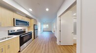 Located at 3417 Harbor Ave SW in Seattle's West Seattle neighborhood, Unit 312 is a thoughtfully designed three-bedroom, two-bathroom residence on a single floor. The open layout flows from the entry directly into the kitchen, which is outfitted with light wood cabinetry, white quartz-style countertops, and a full suite of stainless steel appliances including a refrigerator, electric range, over-the-range microwave, and dishwasher. Warm wood-look laminate flooring runs consistently throughout the unit, creating a cohesive and clean aesthetic.

The living area benefits from natural light streaming through large windows and a glass-panel exterior door that opens to a small outdoor balcony area with views of the surrounding neighborhood. The three bedrooms are compact and efficiently arranged, each featuring the same wood-look flooring and windows that bring in natural light. The primary bedroom spans a cozy footprint ideal for a full or queen bed setup.

Both bathrooms are finished with clean, modern fixtures. One features a full soaking tub with a light blue surround and a large window, while the second includes a stacked washer/dryer unit alongside a vanity and toilet. The unit's overall design is bright, minimal, and move-in ready, making efficient use of every square foot.