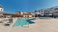 Located at 1000 Banner Drive in Gallatin, Tennessee, this outdoor amenity space is a standout resort-style pool complex designed for relaxation and recreation. The centerpiece is a generously sized swimming pool with crystal-clear water, accompanied by a shallow wading pool featuring in-water lounge chairs — ideal for leisurely sunbathing. A separate spa provides an additional retreat within the complex.

The expansive concrete sun deck surrounds the pools and is furnished with rows of lounge chairs and umbrella-shaded seating areas, offering ample space for residents to unwind under open skies. A charming wood pergola provides a shaded gathering spot, adding architectural warmth to the outdoor environment.

A dedicated poolhouse structure, clad in white siding with stone accent details, serves the pool area with storage and utility access. The entrance to the complex features a sophisticated clubhouse building with natural wood doors, stone masonry, and frameless glass railings, reflecting a modern craftsman aesthetic.

The entire pool area is enclosed by a sleek black iron fence with lush landscaping, including trimmed evergreen shrubs and young trees. The multi-story residential buildings surrounding the complex feature white siding, stone accents, and private balconies, creating a cohesive and upscale community atmosphere beneath the wide Tennessee sky.