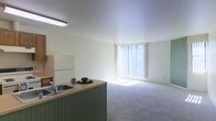 This single-floor unit features an open-concept layout that seamlessly connects the kitchen and living area, creating a cohesive and functional living space. The living area is finished with wall-to-wall carpeting in a neutral tone, contributing to a comfortable and understated atmosphere. A large window dressed with vertical blinds dominates one wall, allowing an abundance of natural light to flood the interior and brighten the overall space.

The kitchen is defined by a green-paneled peninsula with a laminate countertop, which serves as both a workspace and a visual divider between the cooking and living zones. The peninsula features a double-basin sink with a standard faucet. Kitchen cabinetry consists of medium-toned wood upper cabinets, complemented by a white refrigerator and a white gas range with overhead ventilation. Fluorescent lighting fixtures are mounted to the textured ceiling, providing consistent illumination throughout the kitchen area.

The overall design of the space is practical and straightforward, with a clean, neutral color palette of whites and beiges throughout. The open flow between the kitchen and living area enhances the sense of spaciousness within the unit.
