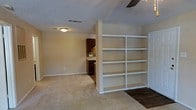 This single-story space offers an open, functional layout connecting the main living area to the kitchen. The living area is finished with neutral carpet and warm beige walls, creating a welcoming atmosphere. A ceiling fan is mounted overhead, complementing the flush-mount lighting. A textured popcorn ceiling adds a classic touch throughout.

A prominent built-in white shelving unit serves as a partial room divider, offering generous display and storage space across multiple shelves. This architectural feature adds both practicality and visual interest while maintaining an open feel between areas.

Transitioning from carpet to hard flooring, the space opens toward the kitchen, which features rich dark cabinetry, a colorful mosaic tile backsplash, and a countertop workspace. The contrast between the dark kitchen finishes and the light tones of the main living area creates a balanced visual dynamic.

Overall, the space delivers a practical and comfortable layout, with thoughtful built-in storage, distinct flooring transitions, and a cohesive neutral color palette that suits a variety of interior styles.