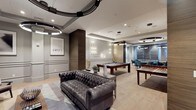 Located at 990 Corporate Drive in Westbury, NY, The Vanderbilt's amenity lounge is a thoughtfully designed social space that combines comfort and style in equal measure. The lounge is anchored by a tufted leather Chesterfield sofa paired with a sleek coffee table, creating an inviting conversation area ideal for relaxing or entertaining. A full-size billiards table serves as a central focal point, positioned beneath contemporary pendant lighting that adds a refined, modern touch. Adjacent to the billiards area, a dedicated game and dining table zone is surrounded by teal upholstered chairs, lending a pop of color to the otherwise neutral palette. The interior design features soft gray wainscoting, warm wood accent panels, and light hardwood-style flooring that together create a cohesive and sophisticated atmosphere. Recessed lighting and elegant wall sconces illuminate the space evenly, while framed artwork contributes a curated, gallery-like quality. The open layout allows each zone to flow naturally into the next, making the lounge equally suited for casual gatherings, friendly competition, or quiet leisure. Overall, the space reflects a polished, upscale aesthetic that elevates the residential amenity experience at The Vanderbilt.