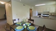 This single-floor residence offers a practical and inviting layout designed to maximize comfort and functionality. The dining area is positioned as a central gathering space, accommodating a four-seat dark wood dining table that anchors the room. The open-concept design creates a seamless flow between the dining area and the adjacent kitchen, making the space feel connected and sociable. The kitchen features a peninsula-style counter with ample workspace, white cabinetry providing generous storage, and a suite of essential appliances including a full-size refrigerator, microwave, and range. Light-toned walls throughout contribute to a bright and airy atmosphere, while overhead lighting ensures the kitchen and dining areas are well-illuminated. A hallway visible from the dining area leads further into the residence, hinting at additional private living spaces beyond. The overall design is clean and straightforward, emphasizing ease of use and a cohesive interior aesthetic. The neutral color palette provides a versatile backdrop, making the space adaptable to a variety of personal styles and furnishing choices. This residence is well-suited for those seeking a manageable, efficiently designed living environment.