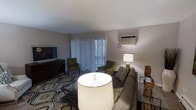 This 1-bedroom, 1-bathroom single-floor residence offers a thoughtfully designed layout that maximizes comfort and functionality. The living area serves as the heart of the space, featuring neutral-toned walls and a soft, light-colored ceiling that creates a clean and airy atmosphere. A large sliding glass door dressed with sheer vertical blinds fills the room with natural light while providing access to the outdoors. The living room accommodates a full seating arrangement, including a plush sofa and accent chairs, complemented by a patterned area rug that adds texture and warmth to the space. A wall-mounted air conditioning unit ensures year-round climate control. A media console with a flat-screen television anchors one side of the room, adding to the space's practical appeal. Decorative lighting, including table lamps, enhances the ambiance and creates a warm, inviting glow. The overall design aesthetic is understated and contemporary, with a neutral color palette that offers a versatile backdrop for personalization. This residence presents a comfortable and well-organized living environment suitable for individuals or couples seeking a practical, move-in-ready home.