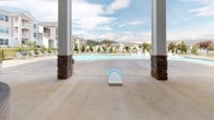 Located at 14 Wooster St in Arden, NC, this single-level outdoor amenity space offers a resort-inspired retreat with a thoughtfully designed covered pavilion. Large stone-accented columns frame the perimeter, supporting a generous roofline outfitted with ceiling fans that provide comfort throughout the warmer months. The centerpiece of the lounge area is a striking floor-to-ceiling stacked stone fireplace, accompanied by a mounted flat-screen television and curved wicker sectional seating upholstered in vibrant teal, creating an inviting gathering space for residents. Multiple seating arrangements are arranged across the smooth concrete deck, offering flexible areas for relaxation and socializing. French doors and floor-to-ceiling windows along the adjacent building façade allow abundant natural light to flow through and provide direct access to interior amenities. Just beyond the covered pavilion, a sparkling resort-style pool extends the outdoor living experience, surrounded by lounge chairs and open to sweeping views of the surrounding Blue Ridge Mountain landscape. The combination of natural stone detailing, neutral tones, and lush greenery gives this space a polished yet welcoming ambiance well-suited for both quiet relaxation and community gatherings.