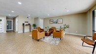 Located in Glendale, Arizona, this single-floor community space offers a thoughtfully designed layout encompassing a living room, dining room, and kitchen. The interior is finished with neutral beige tones throughout, complemented by light tile flooring that runs seamlessly across all areas, lending a clean and cohesive aesthetic.

The spacious living room serves as a central gathering area, furnished with a set of tan leather armchairs arranged around a central coffee table. Desert-themed artwork decorates the walls, nodding to the Arizona locale. A large sliding glass door floods the room with natural light and provides direct access to an outdoor patio area with views of the surrounding grounds.

The dining area features a round wooden table with seating for four and is positioned near another sliding glass door, further enhancing the connection to the outdoors. Adjacent to the dining space is a well-equipped kitchen, highlighted by warm wood cabinetry, dark countertops, a skylight overhead, and black appliances including a full-size refrigerator. A pass-through window connects the kitchen to the neighboring area, promoting an open and communicative flow.

A coffee and beverage station is conveniently situated near the entrance corridor, making it a welcoming feature for visitors and occupants alike. Recessed lighting throughout ensures consistent illumination across all zones of this functional and inviting community space.