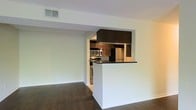 This single-floor residence presents a clean, open-concept layout that seamlessly integrates the living and kitchen areas. The kitchen is a functional centerpiece, featuring rich dark wood cabinetry, dark granite-style countertops, and a full suite of stainless steel appliances, including a refrigerator, microwave, and range. A peninsula counter extends into the living space, providing a natural division between the two areas while maintaining an open and connected feel. The overhead soffit above the kitchen adds a structured architectural detail, framing the space with definition. The living area is generously proportioned, with dark hardwood flooring that runs throughout, lending a warm and cohesive character to the interior. Neutral white walls and a textured ceiling keep the overall palette clean and bright, allowing the darker finishes to serve as deliberate contrast elements. Central air supply vents are integrated into the ceiling, ensuring year-round climate comfort. The space is well-suited for everyday living, offering a practical and straightforward layout that balances open space with purposefully designed functional zones.