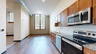 This single-floor unit offers a practical and well-appointed layout spread across a compact footprint, incorporating a kitchen and two bathrooms. The kitchen is a standout feature, equipped with warm-toned wood cabinetry, granite countertops, and a full suite of stainless steel appliances including a refrigerator with ice maker, electric range, built-in microwave, and dishwasher. A classic white subway tile backsplash adds a clean, timeless aesthetic, while dark hardwood-style flooring runs throughout the main living areas, adding warmth and continuity.

The open living space benefits from a tall, multi-pane grid window that draws in generous natural light and lends an industrial-loft character to the unit. Exposed ductwork along the ceiling and a traditional six-panel entry door with transom window above further reinforce this aesthetic. Brick accent walls are visible in areas adjacent to the bathrooms, adding textural contrast to the otherwise smooth, neutral-toned walls.

The bathrooms are functional and neatly finished, each featuring wood-toned vanities with light countertops, a walk-in shower stall, and hardwood-style flooring. Carpeting is present in at least one secondary room area, offering a softer surface underfoot. Overall, the space combines industrial character with modern finishes for a cohesive and livable environment.