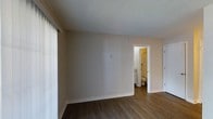 This single-floor space is characterized by its clean, neutral aesthetic and practical layout. The main room features warm-toned hardwood-style flooring that runs throughout, complementing the soft beige walls and white baseboards. A textured popcorn ceiling adds dimension overhead. Vertical blinds along one wall suggest access to natural light through a large window or sliding door. An open doorway leads directly into a bathroom, which is finished in warm-toned lighting and includes a toilet and what appears to be additional fixtures. Adjacent to the bathroom entry, a white door provides access to a closet, offering convenient storage. The overall ambiance of the space is understated and functional, with a neutral color palette that provides a versatile backdrop for a variety of interior styles. The layout is straightforward and efficient, making practical use of the available space throughout.