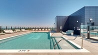Located atop 1860 Reston Row Plaza in Reston, Virginia, this expansive rooftop amenity deck offers a striking blend of modern architecture and open-air leisure. The centerpiece is a generously sized swimming pool with crystal-clear water, complemented by an adjacent spa, both finished in clean, contemporary tile work. Surrounding the pool, large-format stone pavers extend across the entire deck, providing a polished and cohesive aesthetic throughout the space.

Ample seating is arranged poolside in the form of cushioned chaise lounges and daybeds, offering comfortable spots to relax while taking in the surroundings. A covered pergola area with additional lounge seating provides a shaded retreat, furnished with stylish outdoor sofas and accent tables. Glass panel railings line the perimeter of the deck, ensuring unobstructed, panoramic views of the Reston skyline and the lush Virginia landscape stretching to the horizon.

The building's sleek, modern facade of dark-toned metal panels frames the space, reinforcing the sophisticated urban character of the rooftop. With an open sky overhead and far-reaching views on all sides, the deck is bathed in natural light throughout the day. This rooftop destination delivers a resort-caliber experience elevated above the heart of Reston Town Center.