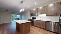 This 1-bedroom, 1-bathroom residence delivers a practical and modern living experience with consistent, quality finishes throughout. The open-concept kitchen is a standout feature, showcasing rich dark-toned cabinetry, granite countertops, a subway tile backsplash, and a full suite of stainless steel appliances including a gas range and built-in microwave. A central kitchen island with pendant lighting above provides additional prep space and serves as a natural gathering point that flows into the living area. The living room is bathed in natural light from a sliding glass door that opens onto a private balcony with leafy outdoor views, and warm hardwood-style flooring runs throughout the main living spaces. The bedroom is a comfortable retreat, featuring plush carpeting, a large window framing serene tree-lined views, and access to a spacious walk-in closet outfitted with wire shelving and hanging systems. The bathroom mirrors the kitchen's design aesthetic with matching dark cabinetry, granite countertops, an undermount sink, and a large vanity mirror. A dedicated in-unit laundry area with a stacked washer and dryer adds everyday convenience, completing this efficiently designed and move-in-ready home.