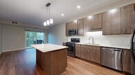 This 1-bedroom, 1-bathroom residence delivers a practical and modern living experience with consistent, quality finishes throughout. The open-concept kitchen is a standout feature, showcasing rich dark-toned cabinetry, granite countertops, a subway tile backsplash, and a full suite of stainless steel appliances including a gas range and built-in microwave. A central kitchen island with pendant lighting above provides additional prep space and serves as a natural gathering point that flows into the living area. The living room is bathed in natural light from a sliding glass door that opens onto a private balcony with leafy outdoor views, and warm hardwood-style flooring runs throughout the main living spaces. The bedroom is a comfortable retreat, featuring plush carpeting, a large window framing serene tree-lined views, and access to a spacious walk-in closet outfitted with wire shelving and hanging systems. The bathroom mirrors the kitchen's design aesthetic with matching dark cabinetry, granite countertops, an undermount sink, and a large vanity mirror. A dedicated in-unit laundry area with a stacked washer and dryer adds everyday convenience, completing this efficiently designed and move-in-ready home.