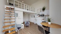 This thoughtfully designed two-floor loft makes clever use of vertical space, with soaring ceilings on the main level that create an airy, open ambiance. The ground floor seamlessly integrates a living area and a compact kitchenette, all finished in a clean, contemporary palette of white cabinetry and warm wood-look flooring. The kitchenette features white upper and lower cabinets, open shelving for display and storage, a microwave, and a smooth white countertop workspace. A round natural wood dining table with modern chairs anchors the dining area, while a seating nook provides a comfortable place to relax. A distinctive white grid-style shelving unit spans the upper mezzanine railing, serving both as a design feature and practical storage visible from below. A sleek spiral staircase with wooden treads connects the two levels. The upper mezzanine level serves as the bedroom, finished with patterned carpet tiles and bathed in natural light from a window. It includes a bed, a small desk area, open shelving, and a ceiling fan. The bathroom is clean and functional, featuring wood-look flooring, a tub-shower combination with a geometric-patterned curtain, a white vanity, and a frosted window for natural light.