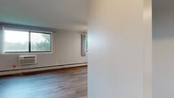 This efficiently designed single-floor unit offers a well-organized layout comprising a combined kitchen and living room, one full bathroom, and a hallway. The living area is bright and open, featuring light hardwood-style flooring, neutral white walls, and baseboard heating. Multiple windows invite natural light and offer leafy outdoor views, enhancing the airy feel of the space. A ceiling fan adds both function and a subtle design element to the living room, which also includes built-in closet storage.

The kitchen is thoughtfully appointed with rich, dark wood cabinetry, stainless steel appliances including a gas range and refrigerator, a subway tile backsplash, a wall-mounted range hood, and light stone countertops. The kitchen flows naturally into the living area, creating a cohesive open-concept feel.

The bathroom is clean and functional, finished with light marble-style tile flooring and a full tub-shower combination in a crisp all-white surround. The hallway connects the spaces efficiently, contributing to the unit's practical flow. Overall, the unit presents a tidy, modern aesthetic with durable finishes well-suited for comfortable everyday living.