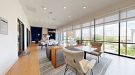 Located at 1614 Hill St in Grand Prairie, Texas, this thoughtfully designed communal space offers a sophisticated and functional environment across a single floor. The open-plan layout is anchored by an expansive lounge area featuring a mix of seating options, including channeled-leather sofas, upholstered armchairs, and sleek dining tables with modern chairs, all arranged on layered area rugs over light hardwood flooring. Bold navy blue textured accent walls create dramatic focal points throughout, contrasting beautifully with the bright, neutral tones of the ceiling and surrounding walls. Floor-to-ceiling windows line the perimeter, flooding the interior with abundant natural light and offering open views of the surrounding outdoor landscape. Sheer and roller shade window treatments allow for flexible light control. A built-in counter-height workspace with orange swivel chairs provides a dedicated area for focused work or collaboration. Recessed lighting and pendant fixtures complement the natural light, maintaining a warm and inviting ambiance. Glass-partitioned private rooms with sleek black frames offer quieter spaces for meetings or individual use, each equipped with wall-mounted displays. A small kitchenette with stone countertops and cabinetry adds convenience. The overall design balances contemporary aesthetics with practical functionality, creating a versatile and welcoming environment.
