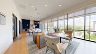 Located at 1614 Hill St in Grand Prairie, Texas, this thoughtfully designed communal space offers a sophisticated and functional environment across a single floor. The open-plan layout is anchored by an expansive lounge area featuring a mix of seating options, including channeled-leather sofas, upholstered armchairs, and sleek dining tables with modern chairs, all arranged on layered area rugs over light hardwood flooring. Bold navy blue textured accent walls create dramatic focal points throughout, contrasting beautifully with the bright, neutral tones of the ceiling and surrounding walls. Floor-to-ceiling windows line the perimeter, flooding the interior with abundant natural light and offering open views of the surrounding outdoor landscape. Sheer and roller shade window treatments allow for flexible light control. A built-in counter-height workspace with orange swivel chairs provides a dedicated area for focused work or collaboration. Recessed lighting and pendant fixtures complement the natural light, maintaining a warm and inviting ambiance. Glass-partitioned private rooms with sleek black frames offer quieter spaces for meetings or individual use, each equipped with wall-mounted displays. A small kitchenette with stone countertops and cabinetry adds convenience. The overall design balances contemporary aesthetics with practical functionality, creating a versatile and welcoming environment.