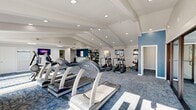 Located at 3131 Mountain Creek Road in Chattanooga, Tennessee, this single-floor fitness center offers a generously sized, thoughtfully designed workout facility. The space is defined by striking vaulted ceilings with exposed white beams and abundant recessed lighting, creating a bright and energizing atmosphere throughout.

The main workout area is anchored by a row of True-brand treadmills and is complemented by a variety of cardio equipment, including elliptical trainers, stationary bikes, and a rowing machine. A dedicated free weights and functional training zone features a wall-mounted dumbbell and medicine ball rack, stability balls, and kettlebells, providing options for diverse training routines. Flat-screen televisions are mounted at multiple points throughout the space to keep members entertained during workouts.

A glass-walled studio room, framed by sleek black-trimmed partitions, offers a semi-private area suitable for stretching, yoga, or personal training sessions. The bold blue accent walls add a modern, energetic design element that contrasts cleanly with the white ceilings and light gray walls. Patterned blue-toned carpeting runs throughout, contributing both comfort and contemporary style.

Natural light filters in through several windows, and a small service counter with storage is positioned near the rear of the space. The overall layout is open and efficient, supporting a full range of fitness activities in a clean, modern environment.